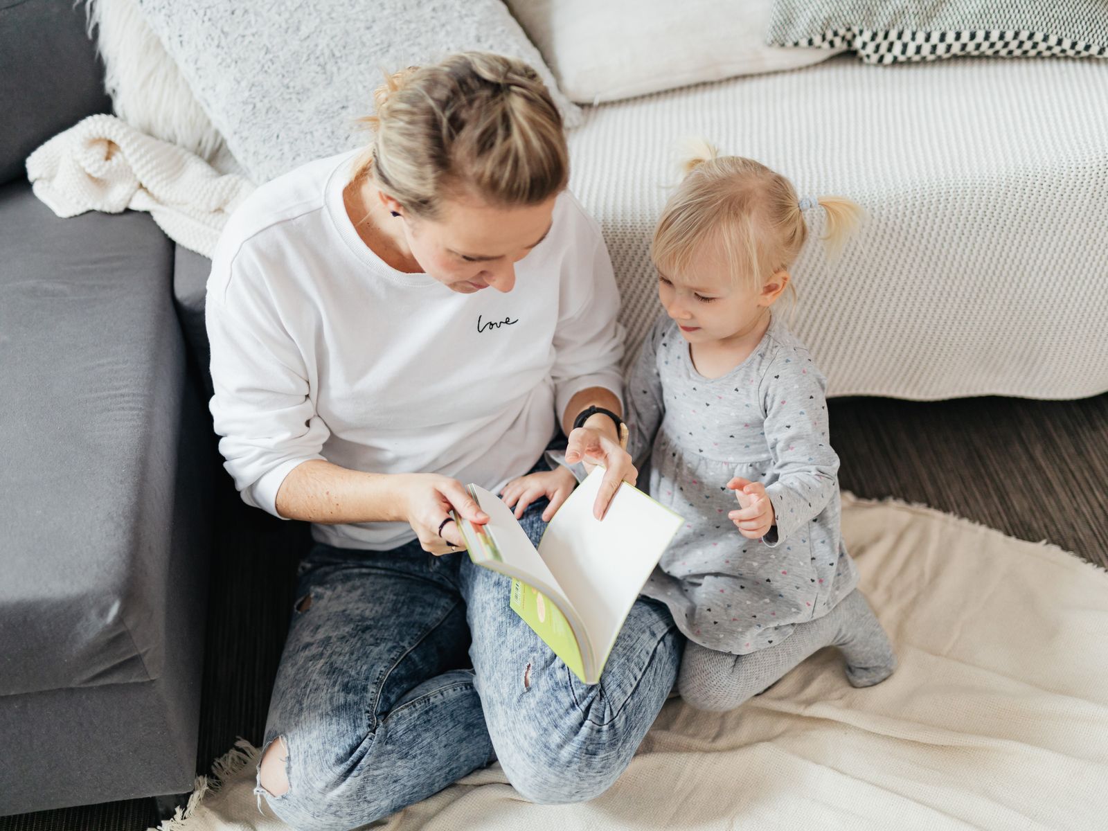 A mother and her toddler daughter reading a picture book together on the couch at home, sharing a warm moment between routine and learning