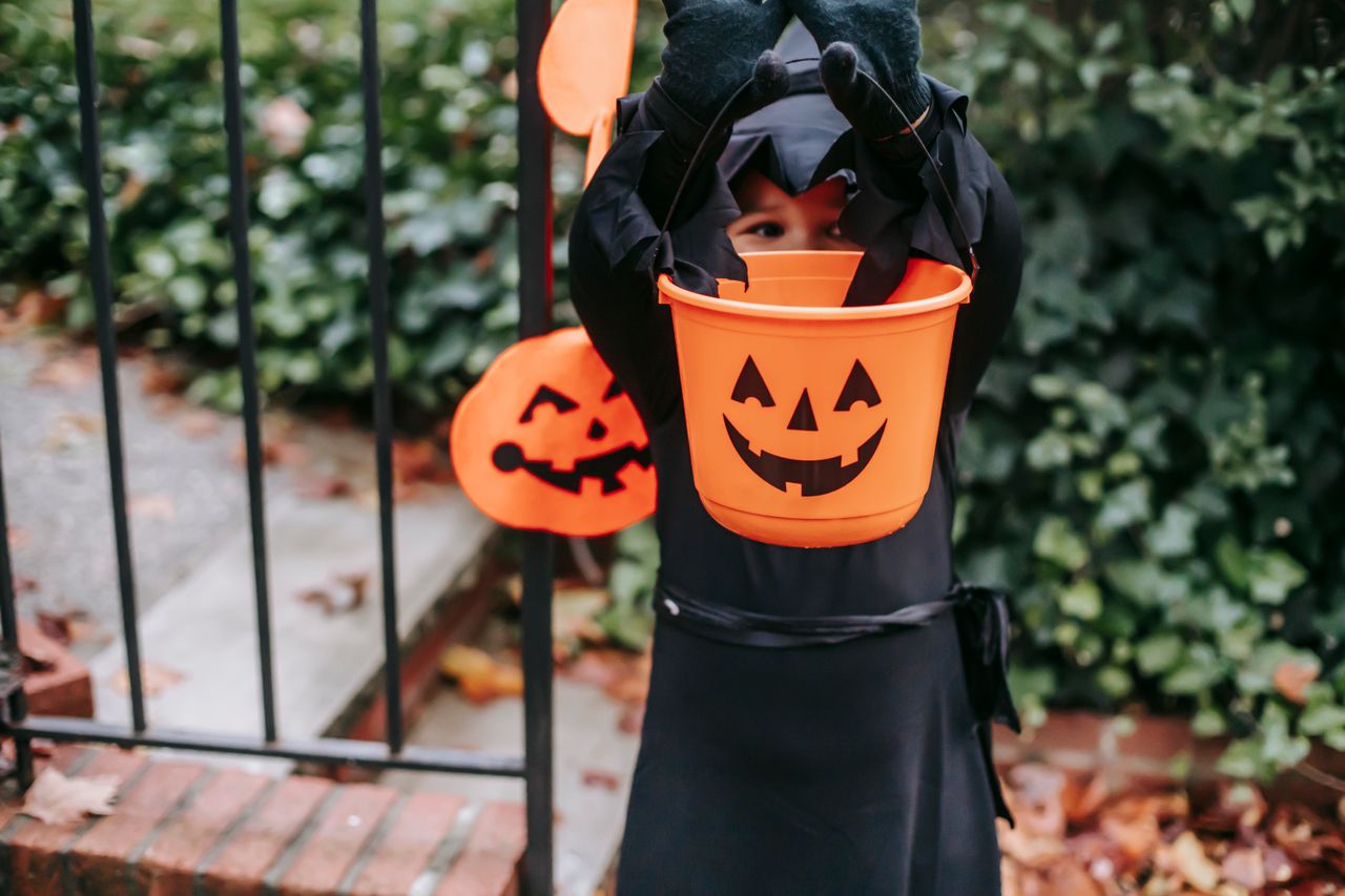 A child dressed in a black costume holds an orange pumpkin bucket outdoors for Halloween